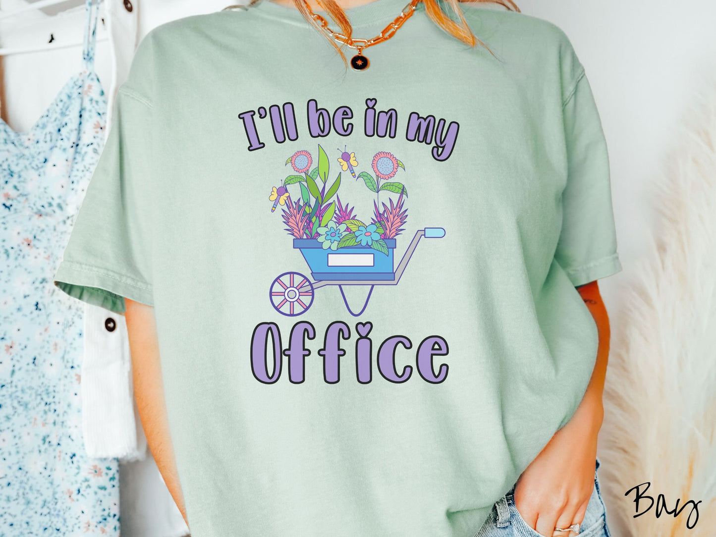 A woman wearing a cute, vintage bay colored Comfort Colors t-shirt with the text I’ll Be in My Office in purple font across the front. In between the text is a blue wheelbarrow with a pink wheel stacked full of colorful plants and flowers.