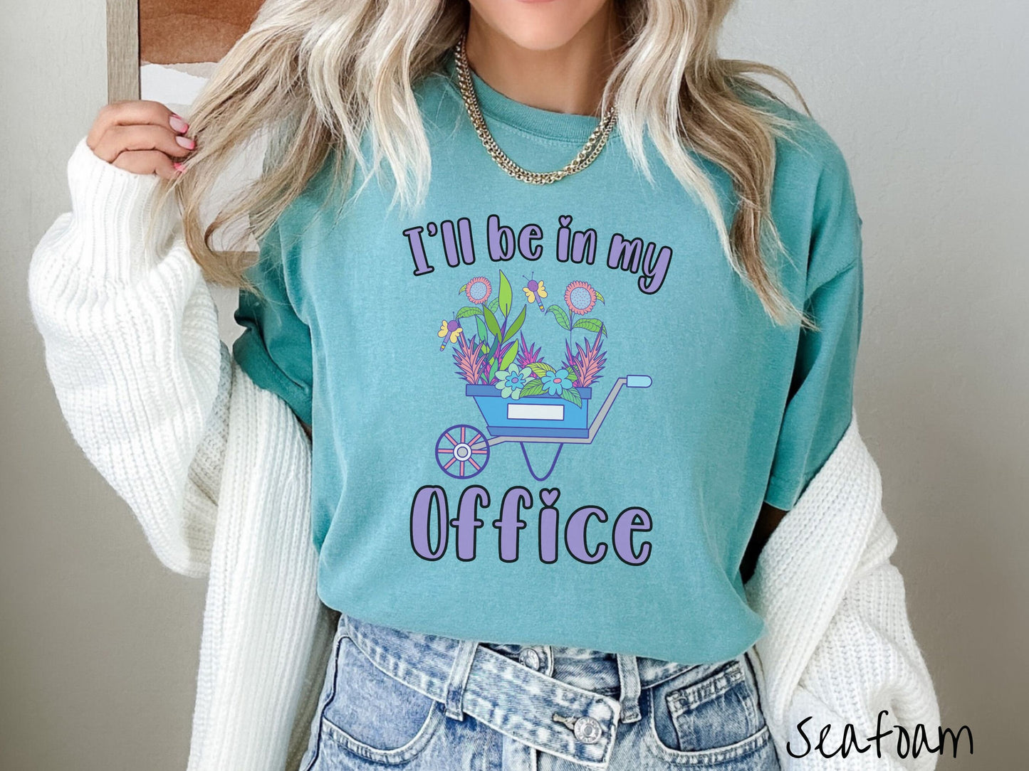 A woman wearing a cute, vintage seafoam colored Comfort Colors t-shirt with the text I’ll Be in My Office in purple font across the front. In between the text is a blue wheelbarrow with a pink wheel stacked full of colorful plants and flowers.