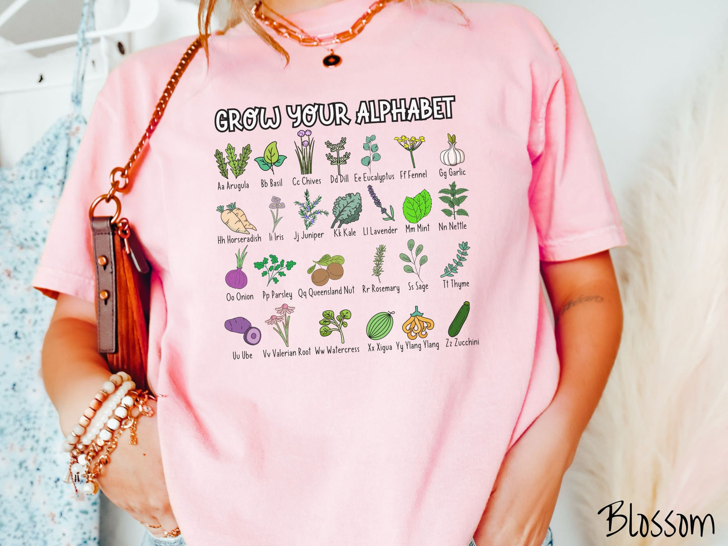 A woman wearing a cute, vintage blossom colored Comfort Colors t-shirt with the text Grow Your Alphabet across the top. Below this is a 4 x 7 grid of different kinds of plants and their names in alphabetical order like Arugula, Basil, and Chives.
