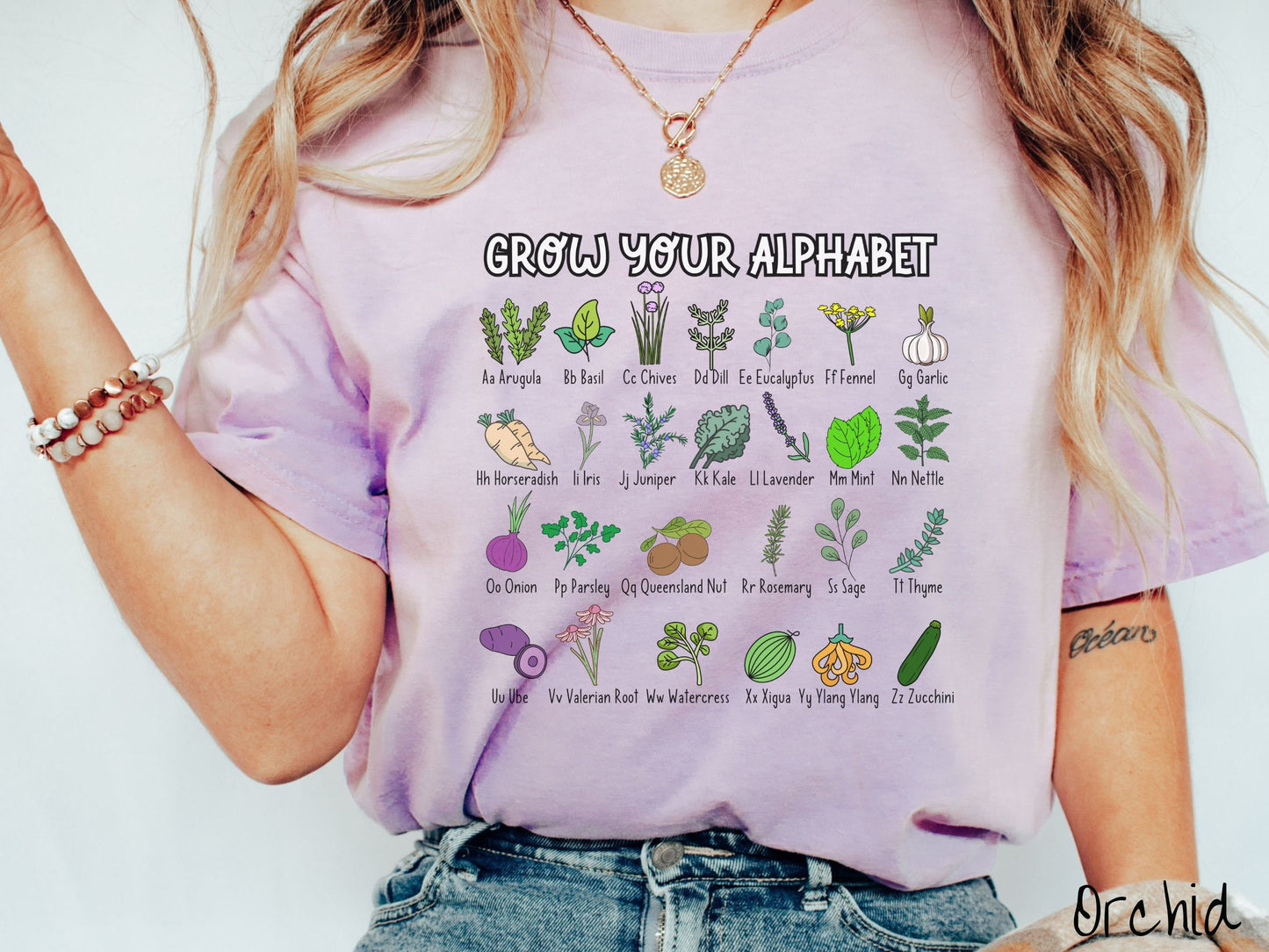 A woman wearing a cute, vintage orchid colored Comfort Colors t-shirt with the text Grow Your Alphabet across the top. Below this is a 4 x 7 grid of different kinds of plants and their names in alphabetical order like Arugula, Basil, and Chives.