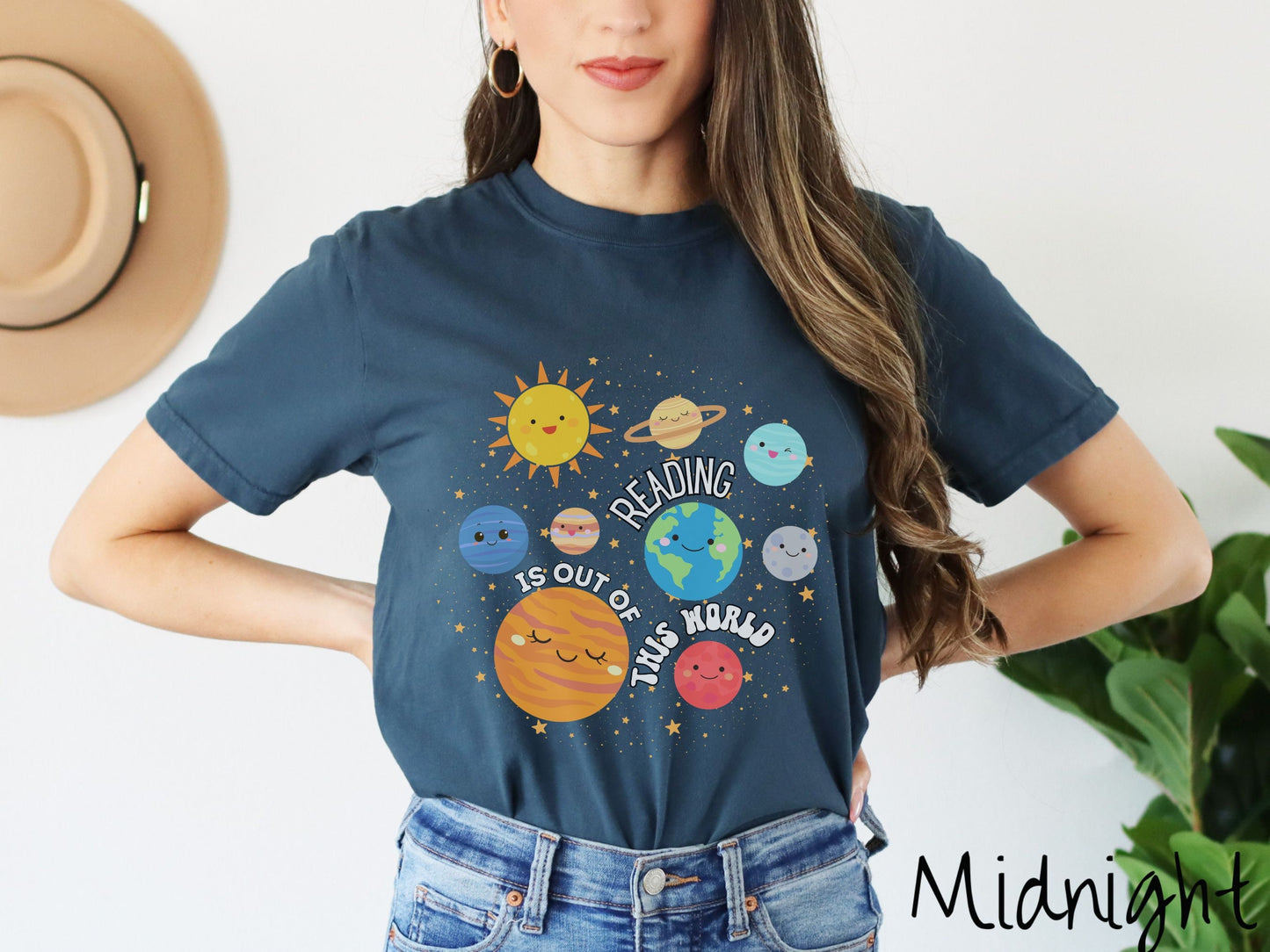 A woman wearing a cute, vintage midnight colored shirt showing the solar system and all the planets have smiling, happy faces. There is text saying Reading Is Out of This World among the planets, and there are many stars twinkling in the background.