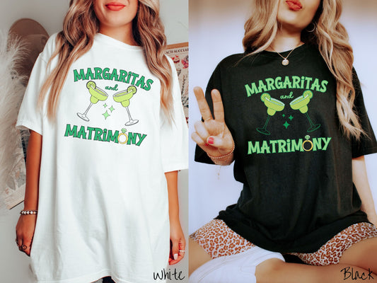 Two women wearing cute, vintage white and black colored Comfort Colors t-shirts with text Margaritas and Matrimony in pink font with two green margarita glasses toasting, each with salt and limes. The O in Matrimony is a diamond wedding ring.