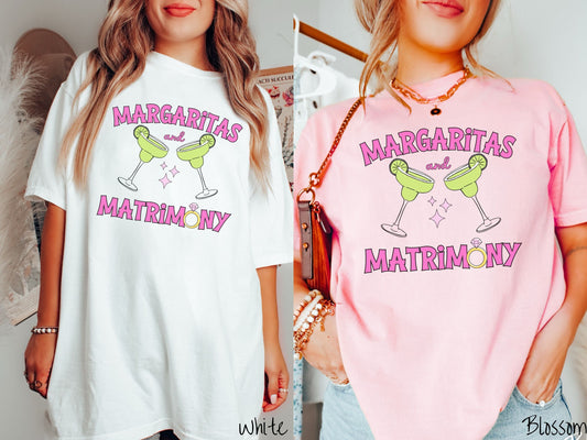 Two women wearing cute, vintage white and blossom colored Comfort Colors t-shirts with text Margaritas and Matrimony in pink font with two green margarita glasses toasting, each with salt and limes. The O in Matrimony is a diamond wedding ring.