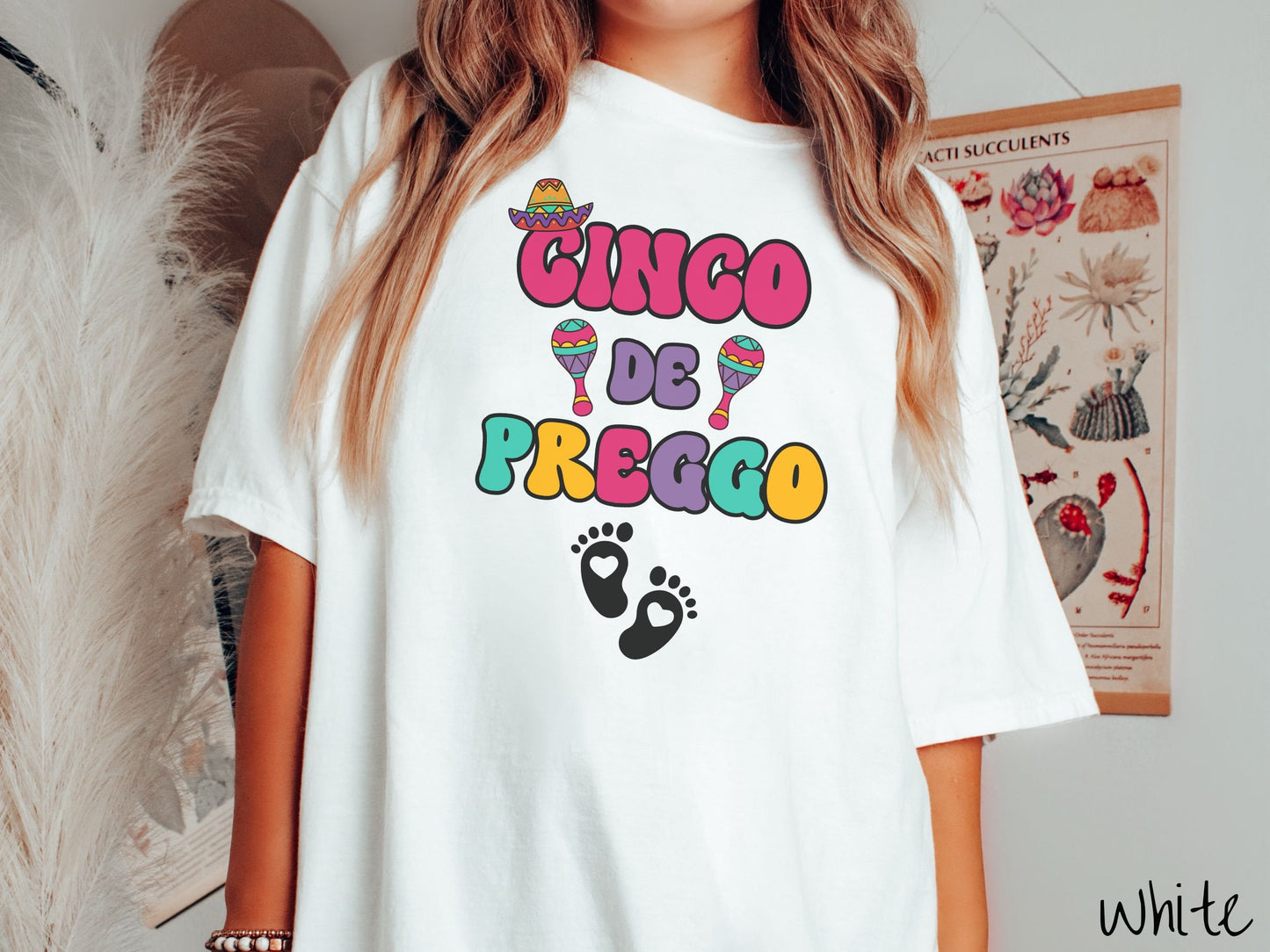 A woman wearing a cute, vintage white colored Comfort Colors t-shirt with the text Cinco de Preggo in colorful font. A sombrero is hanging on top of the C in Cinco, colorful maracas around the word de, and two baby footprints with hearts inside.