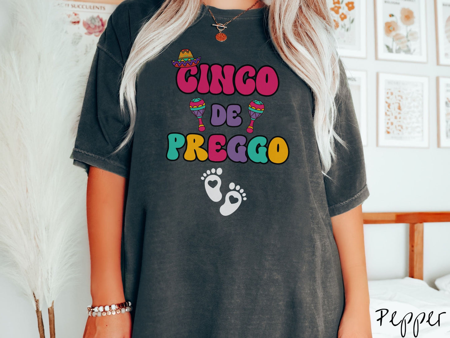 A woman wearing a cute, vintage pepper colored Comfort Colors t-shirt with the text Cinco de Preggo in colorful font. A sombrero is hanging on top of the C in Cinco, colorful maracas around the word de, and two baby footprints with hearts inside.