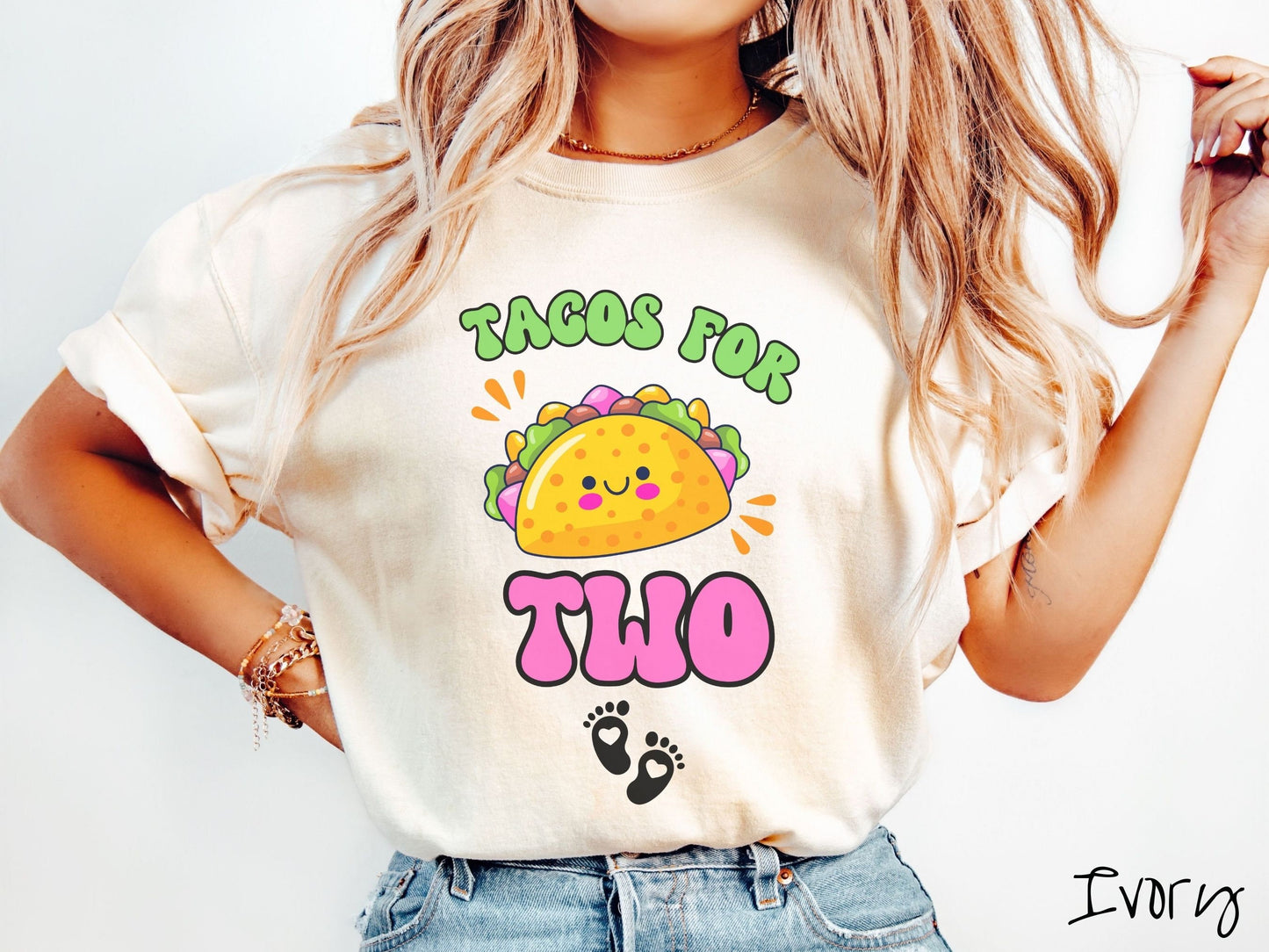A woman wearing a cute, vintage ivory colored Comfort Colors t-shirt with text Tacos for Two in green and pink font. In between the font is a smiling taco with colorful fillings and below are two baby footprints with hearts inside.