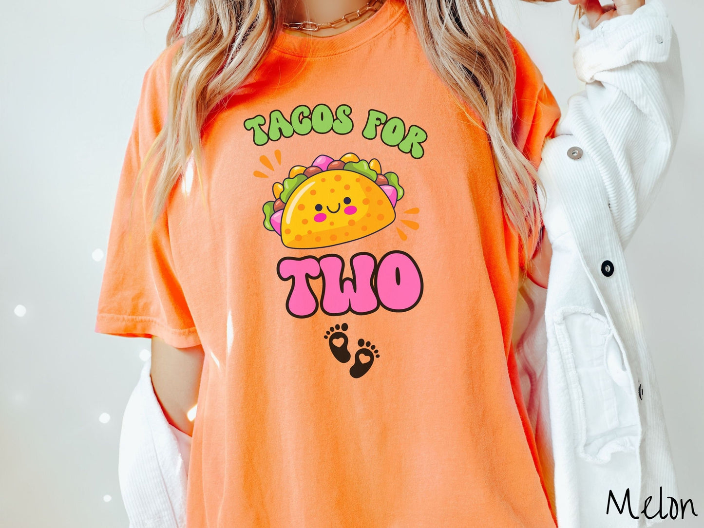 A woman wearing a cute, vintage melon colored Comfort Colors t-shirt with text Tacos for Two in green and pink font. In between the font is a smiling taco with colorful fillings and below are two baby footprints with hearts inside.