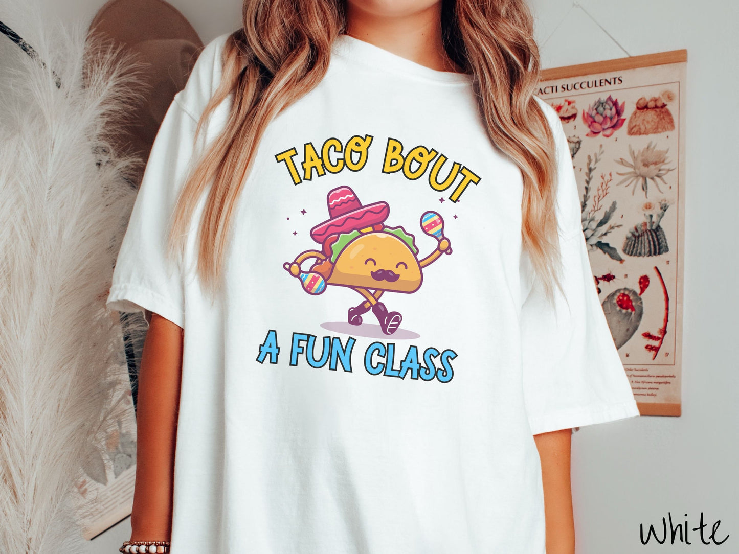 A woman wearing a cute, vintage white colored Comfort Colors t-shirt with text Taco Bout a Fun Class in yellow and blue font. In between the text is a smiling taco with a black mustache shaking colorful tambourines and wearing a red sombrero.