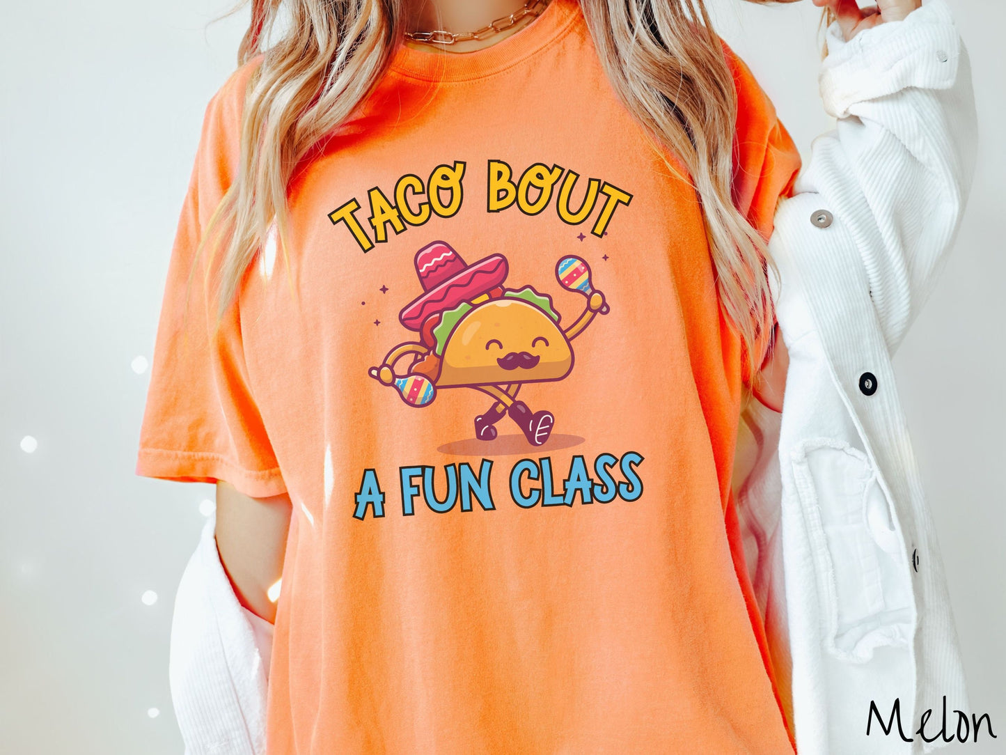 A woman wearing a cute, vintage melon colored Comfort Colors t-shirt with text Taco Bout a Fun Class in yellow and blue font. In between the text is a smiling taco with a black mustache shaking colorful tambourines and wearing a red sombrero.
