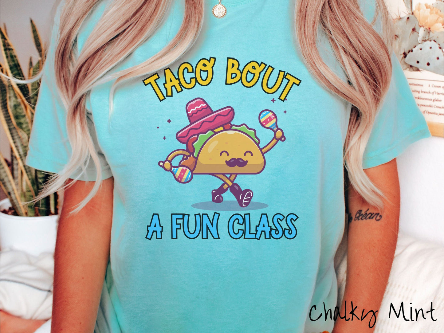 A woman wearing a cute, vintage chalky mint colored Comfort Colors t-shirt with text Taco Bout a Fun Class in yellow and blue font. In between the text is a smiling taco with a black mustache shaking colorful tambourines and wearing a red sombrero.