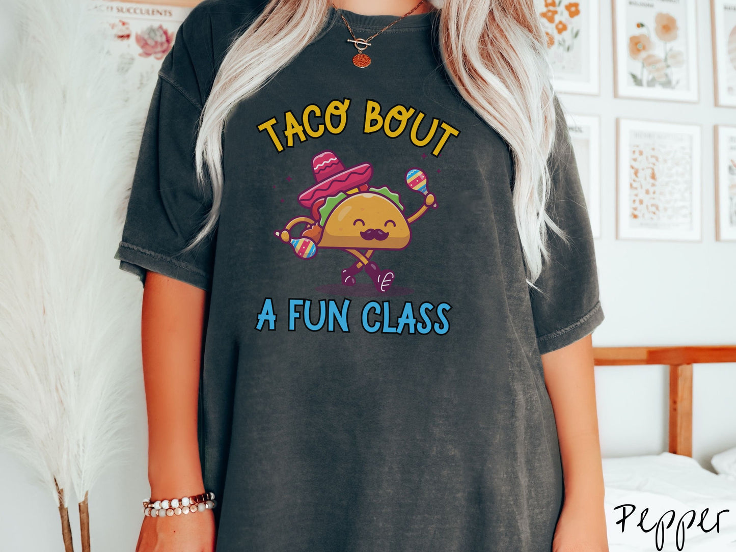 A woman wearing a cute, vintage pepper colored Comfort Colors t-shirt with text Taco Bout a Fun Class in yellow and blue font. In between the text is a smiling taco with a black mustache shaking colorful tambourines and wearing a red sombrero.