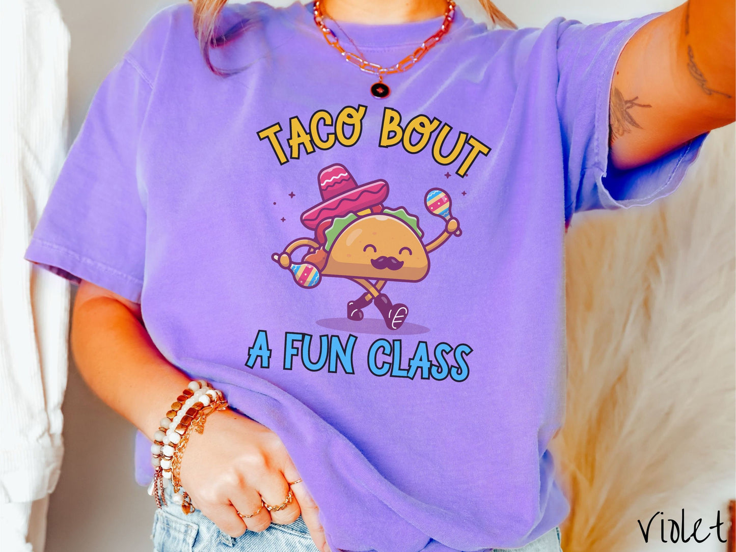 A woman wearing a cute, vintage violet colored Comfort Colors t-shirt with text Taco Bout a Fun Class in yellow and blue font. In between the text is a smiling taco with a black mustache shaking colorful tambourines and wearing a red sombrero.
