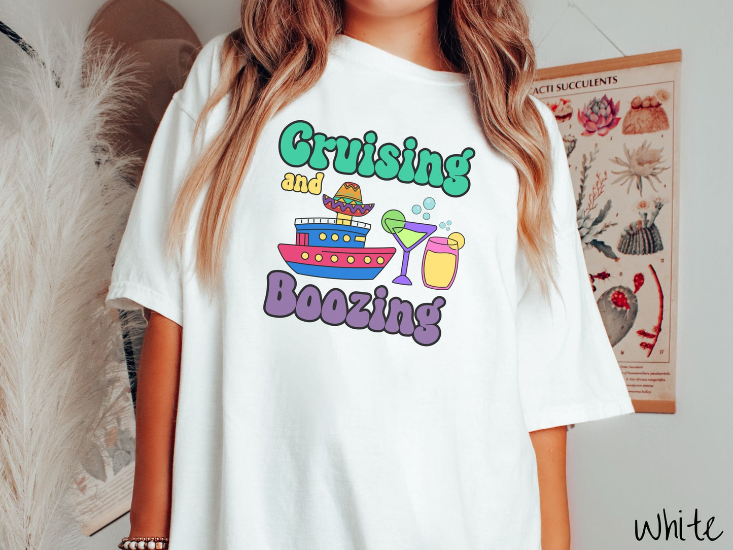 A woman wearing a cute, vintage white colored Comfort Colors t-shirt with text Cruising and Boozing in green, yellow, and purple font. Between the text are a colorful cruise ship wearing a sombrero and two cocktail glasses with margaritas and limes.