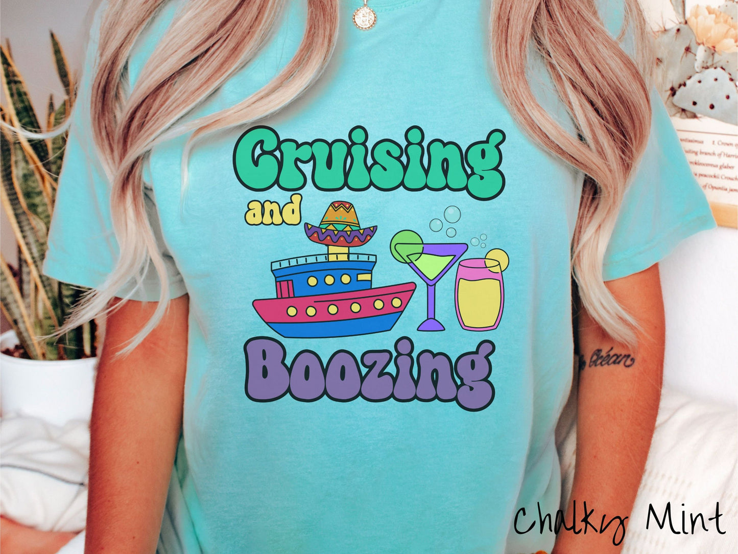 A woman wearing a cute, vintage chalky mint colored Comfort Colors t-shirt with text Cruising and Boozing in green, yellow, and purple font. Between the text are a cruise ship wearing a sombrero and two cocktail glasses with margaritas and limes.