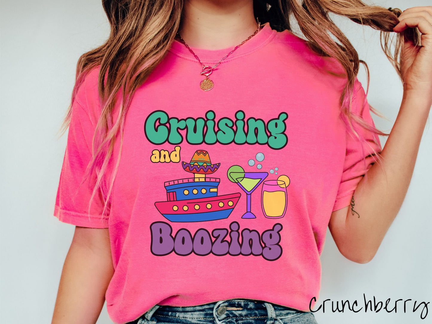 A woman wearing a cute, vintage crunchberry colored Comfort Colors t-shirt with text Cruising and Boozing in green, yellow, and purple font. Between the text are a cruise ship wearing a sombrero and two cocktail glasses with margaritas and limes.