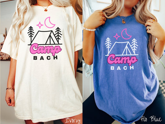 Two women wearing cute, vintage ivory and flo blue colored Comfort Colors t-shirts with text Camp Bach in pink and black font. Above the text is an outline of a tent in between two pine trees with a pink star and moon above in the sky.