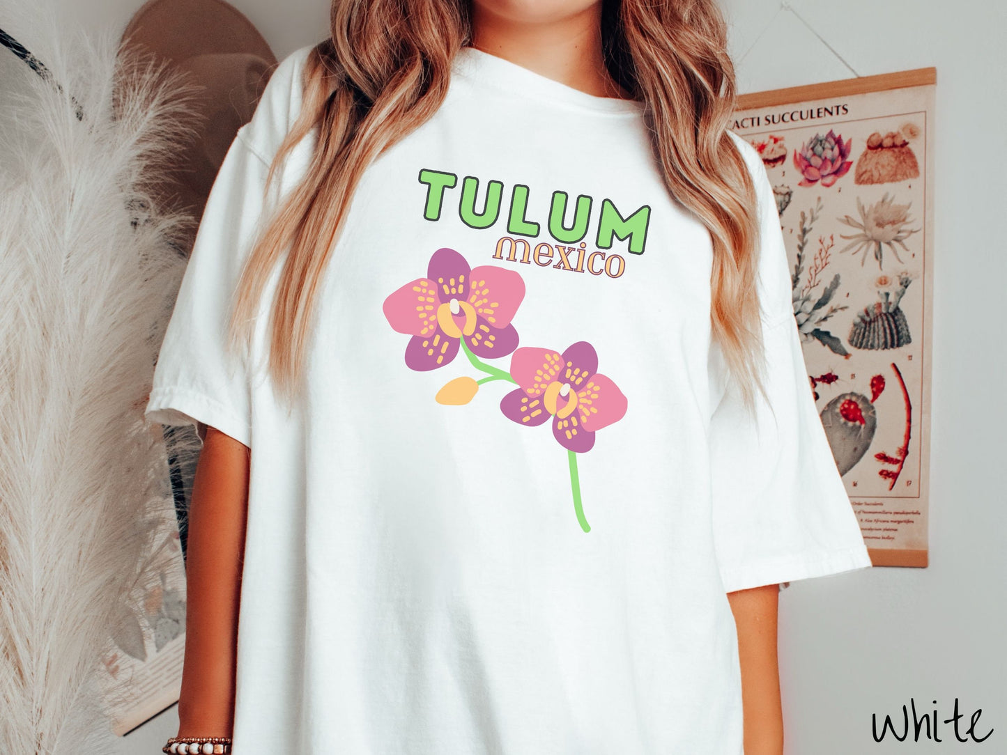 A woman wearing a vintage, white colored shirt with the text Tulum Mexico in green and yellow font, respectively, at the top and below it are two beautiful pink and purple flowers with green stems.