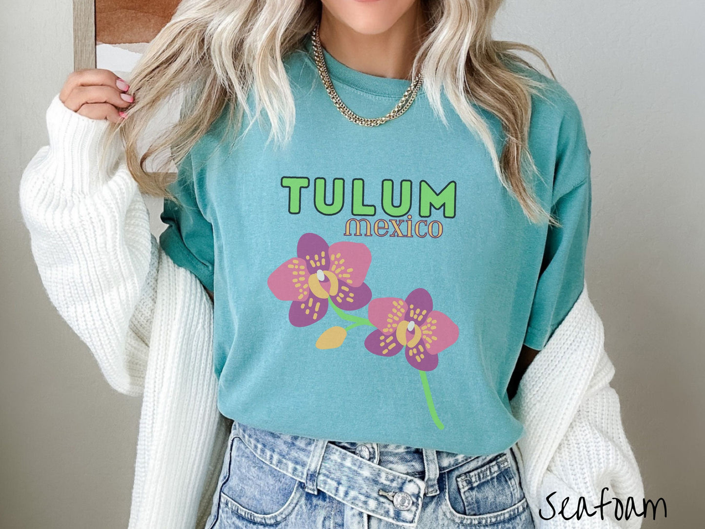 A woman wearing a vintage, seafoam colored shirt with the text Tulum Mexico in green and yellow font, respectively, at the top and below it are two beautiful pink and purple flowers with green stems.