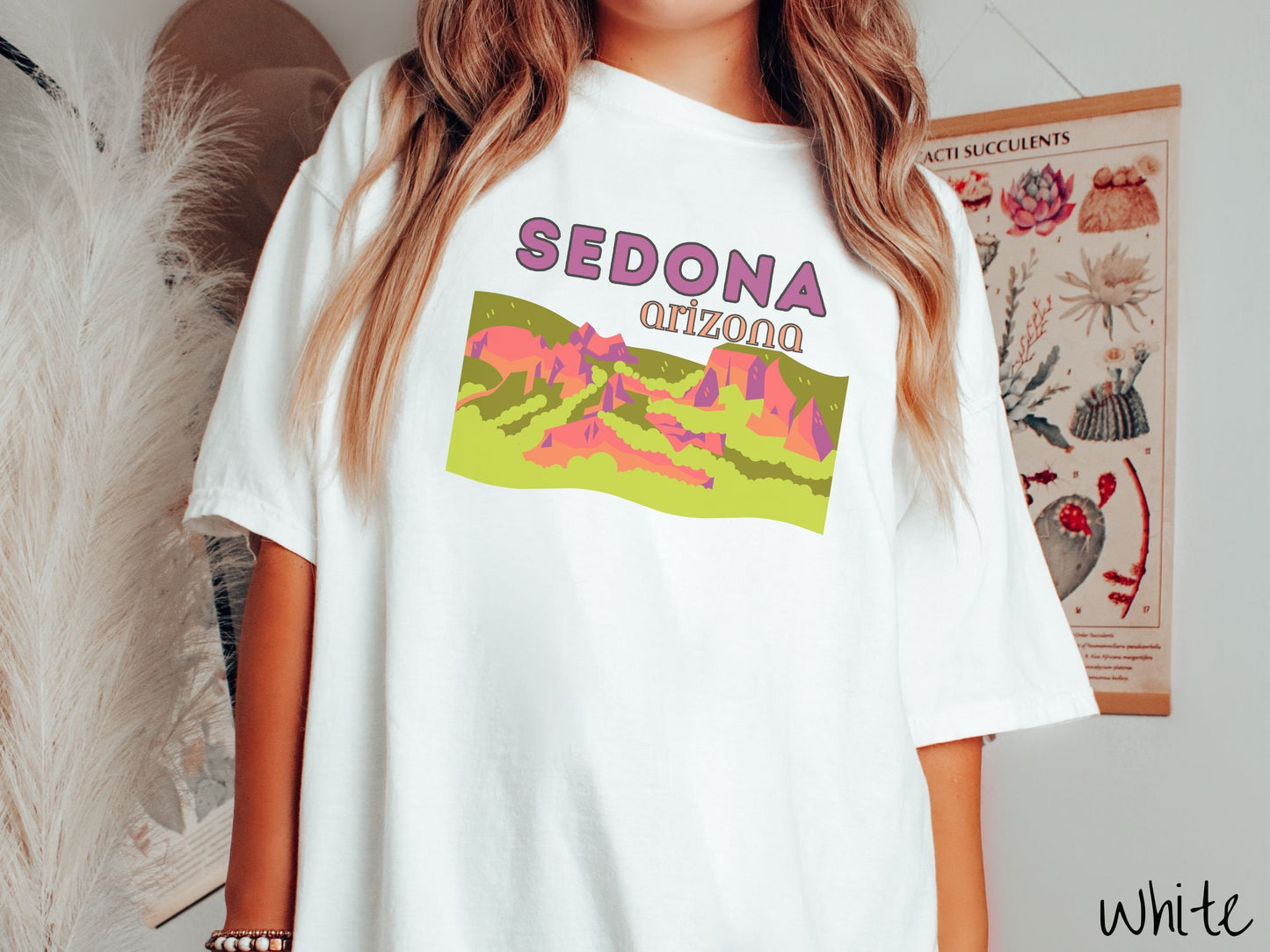 A woman wearing a vintage, white colored shirt with the text Sedona Arizona in purple and orange font, respectively, and below that is a picture of red rock canyons and green meadows.