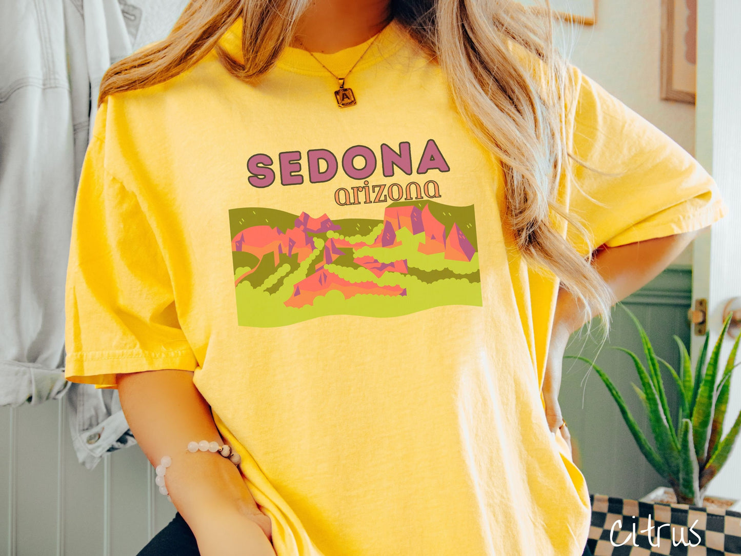A woman wearing a vintage, citrus colored shirt with the text Sedona Arizona in purple and orange font, respectively, and below that is a picture of red rock canyons and green meadows.