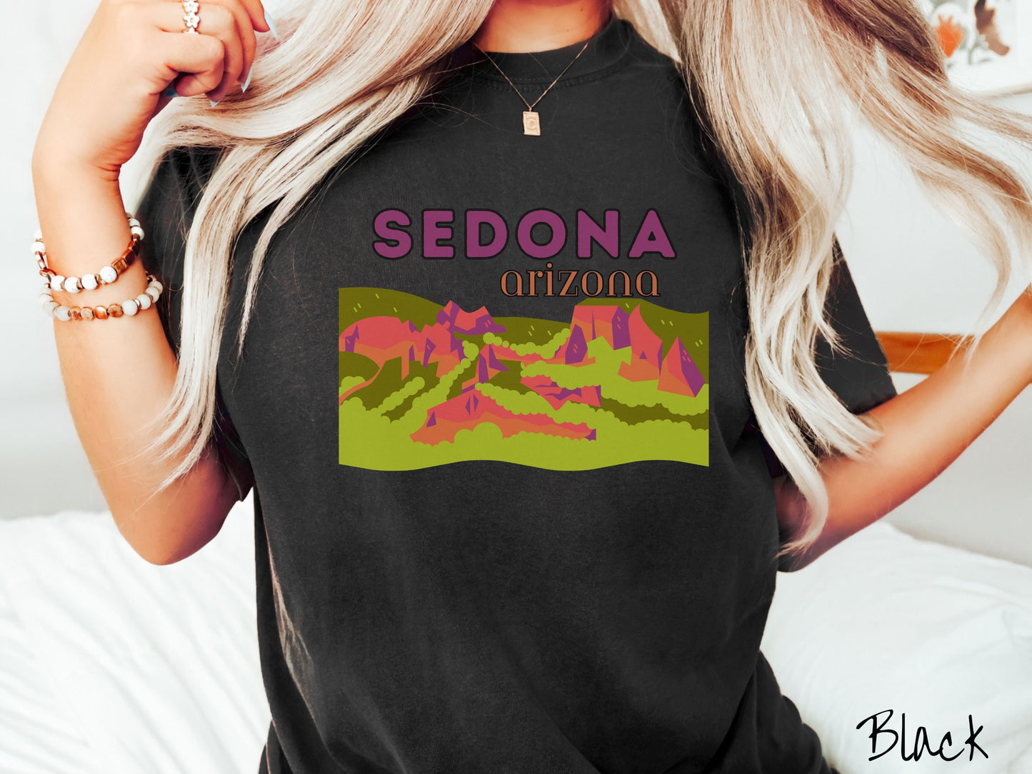 A woman wearing a vintage, black colored shirt with the text Sedona Arizona in purple and orange font, respectively, and below that is a picture of red rock canyons and green meadows.