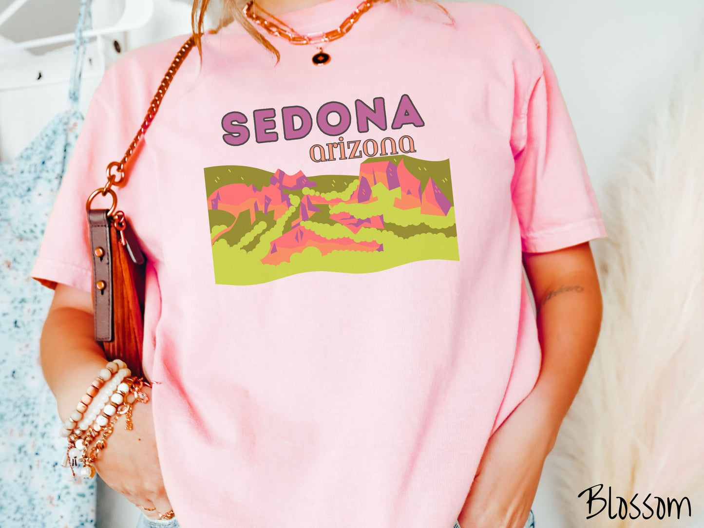 A woman wearing a vintage, blossom colored shirt with the text Sedona Arizona in purple and orange font, respectively, and below that is a picture of red rock canyons and green meadows.