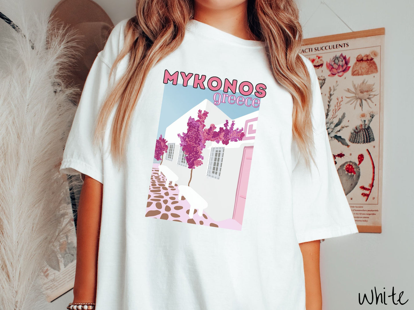 A woman wearing a vintage, white colored shirt with the text Mykonos Greece at the top, and beneath is a picture of Cycladic architecture, a beautiful white home with purple trees in the front yard, and a pink driveway with brown stone.