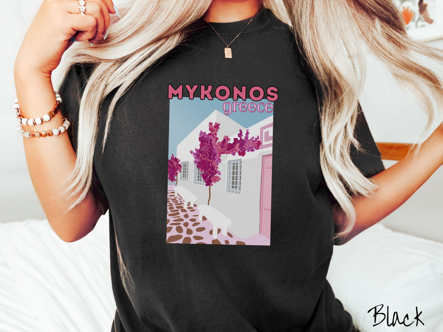 A woman wearing a vintage, black colored shirt with the text Mykonos Greece at the top, and beneath is a picture of Cycladic architecture, a beautiful white home with purple trees in the front yard, and a pink driveway with brown stone.
