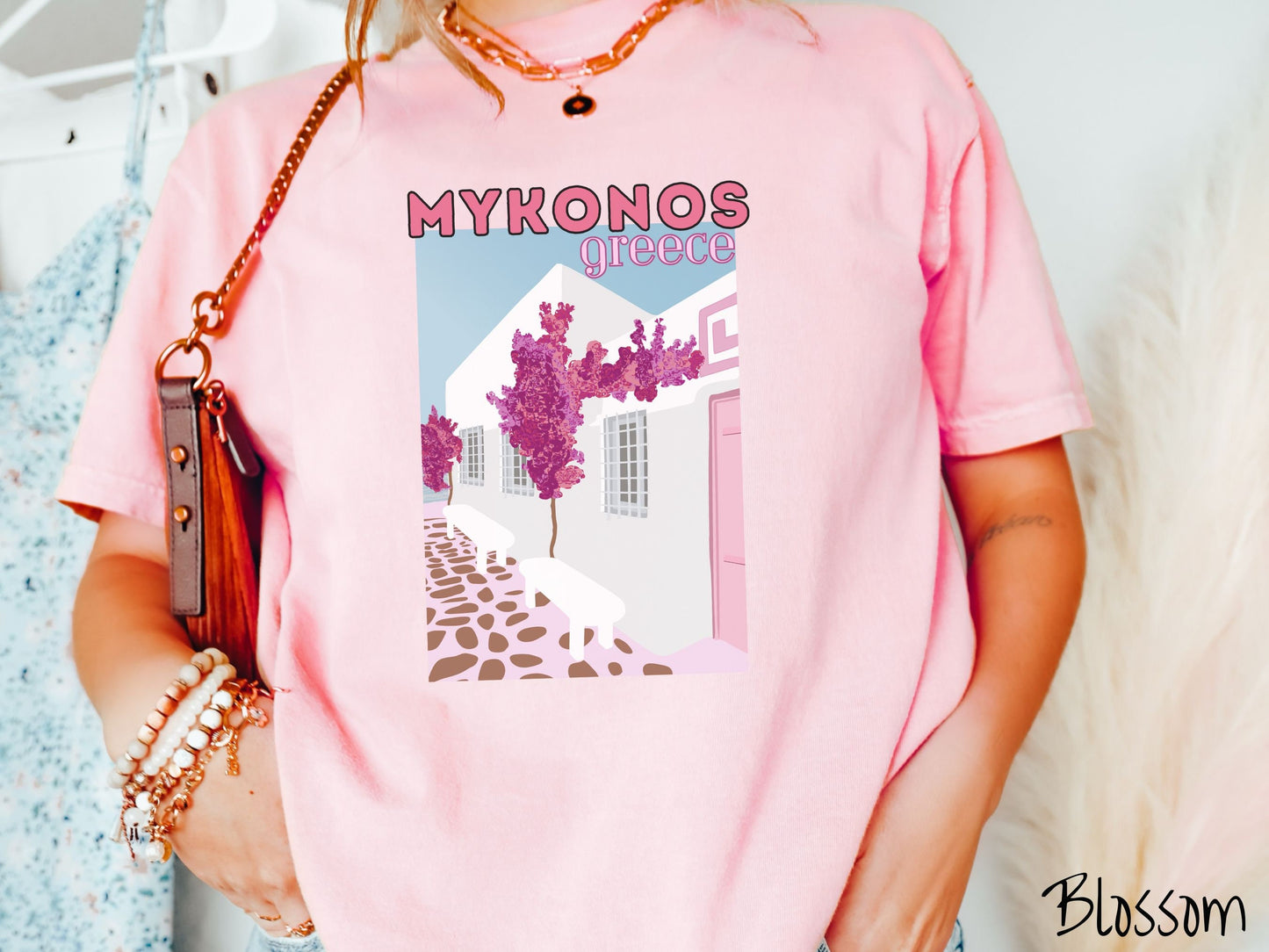 A woman wearing a vintage, blossom colored shirt with the text Mykonos Greece at the top, and beneath is a picture of Cycladic architecture, a beautiful white home with purple trees in the front yard, and a pink driveway with brown stone.