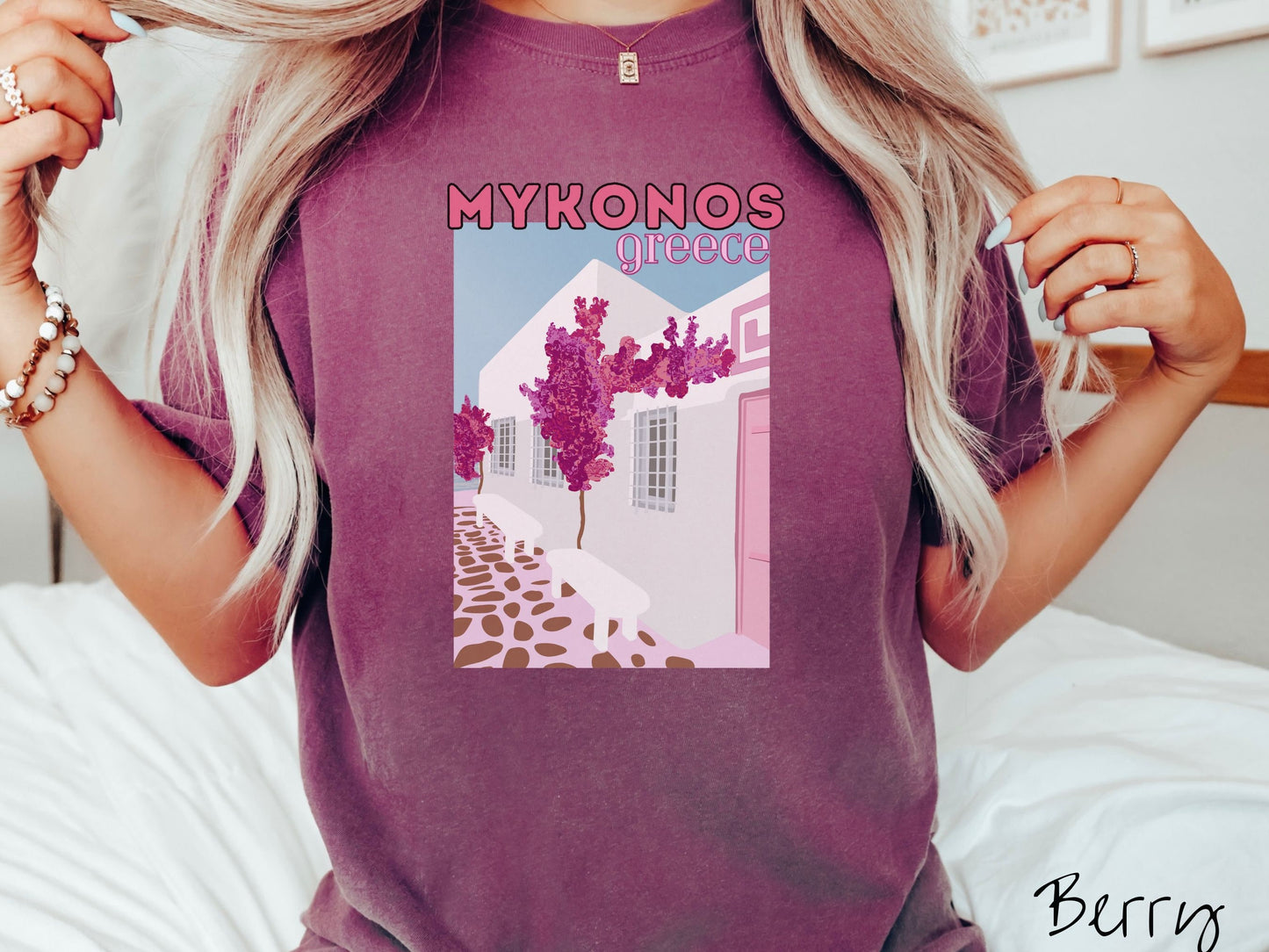 A woman wearing a vintage, berry colored shirt with the text Mykonos Greece at the top, and beneath is a picture of Cycladic architecture, a beautiful white home with purple trees in the front yard, and a pink driveway with brown stone.