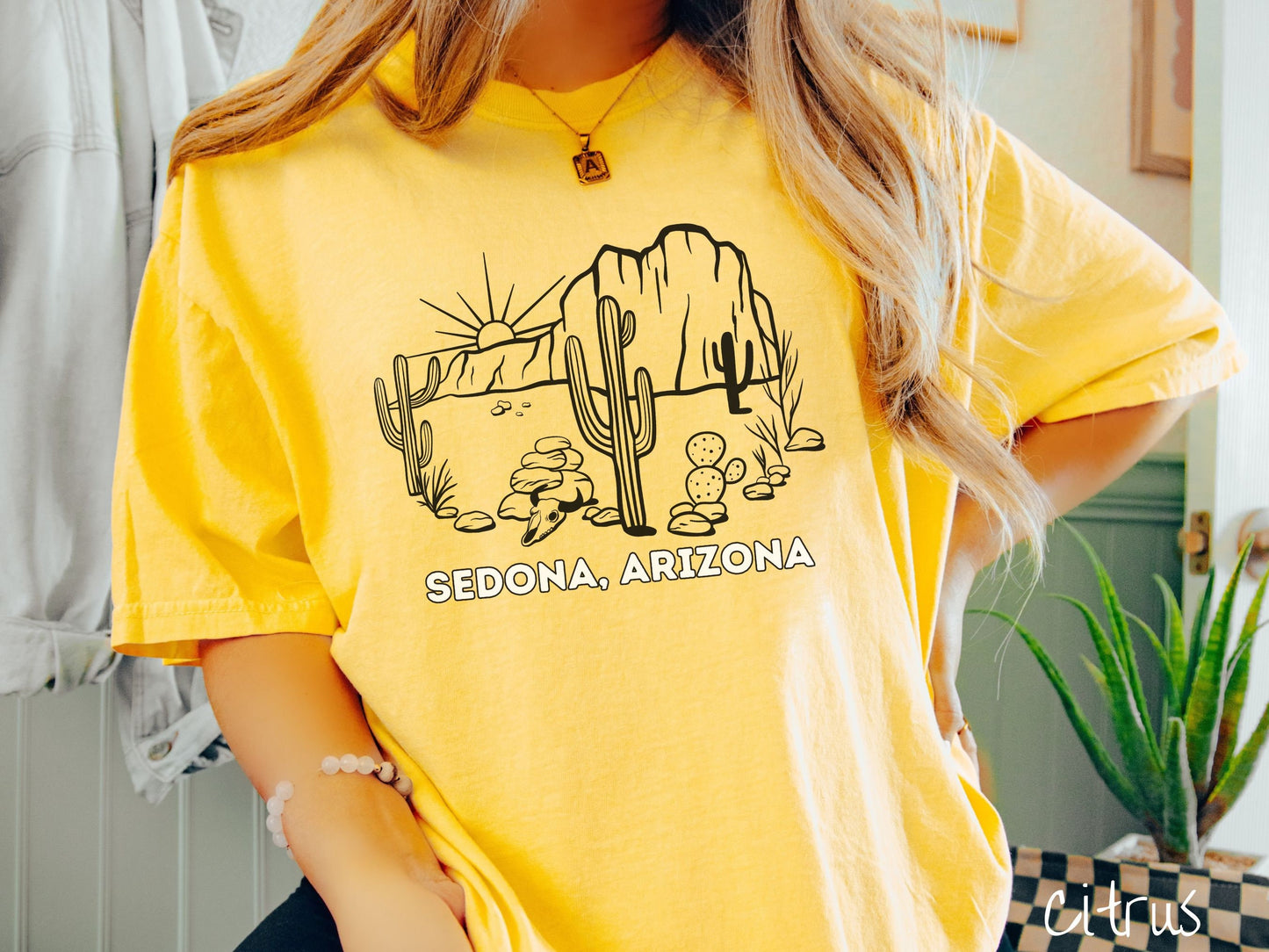 A woman wearing a cute, vintage citrus colored Comfort Colors t-shirt with text Sedona, Arizona in white font. Above is a black outline of a desert landscape with the sun and mountains in the background, and cacti and rocks in the foreground.