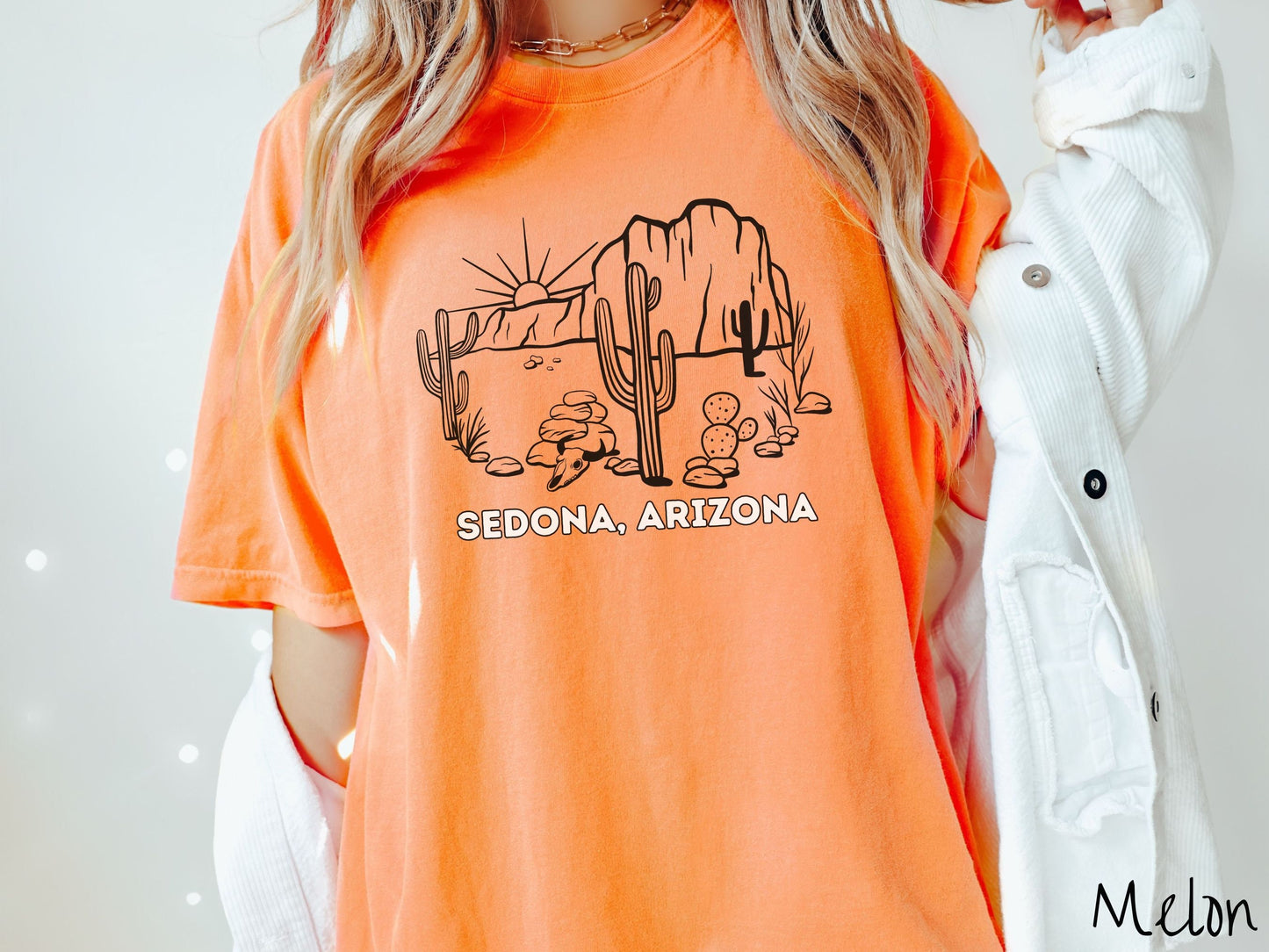 A woman wearing a cute, vintage melon colored Comfort Colors t-shirt with text Sedona, Arizona in white font. Above is a black outline of a desert landscape with the sun and mountains in the background, and cacti and rocks in the foreground.