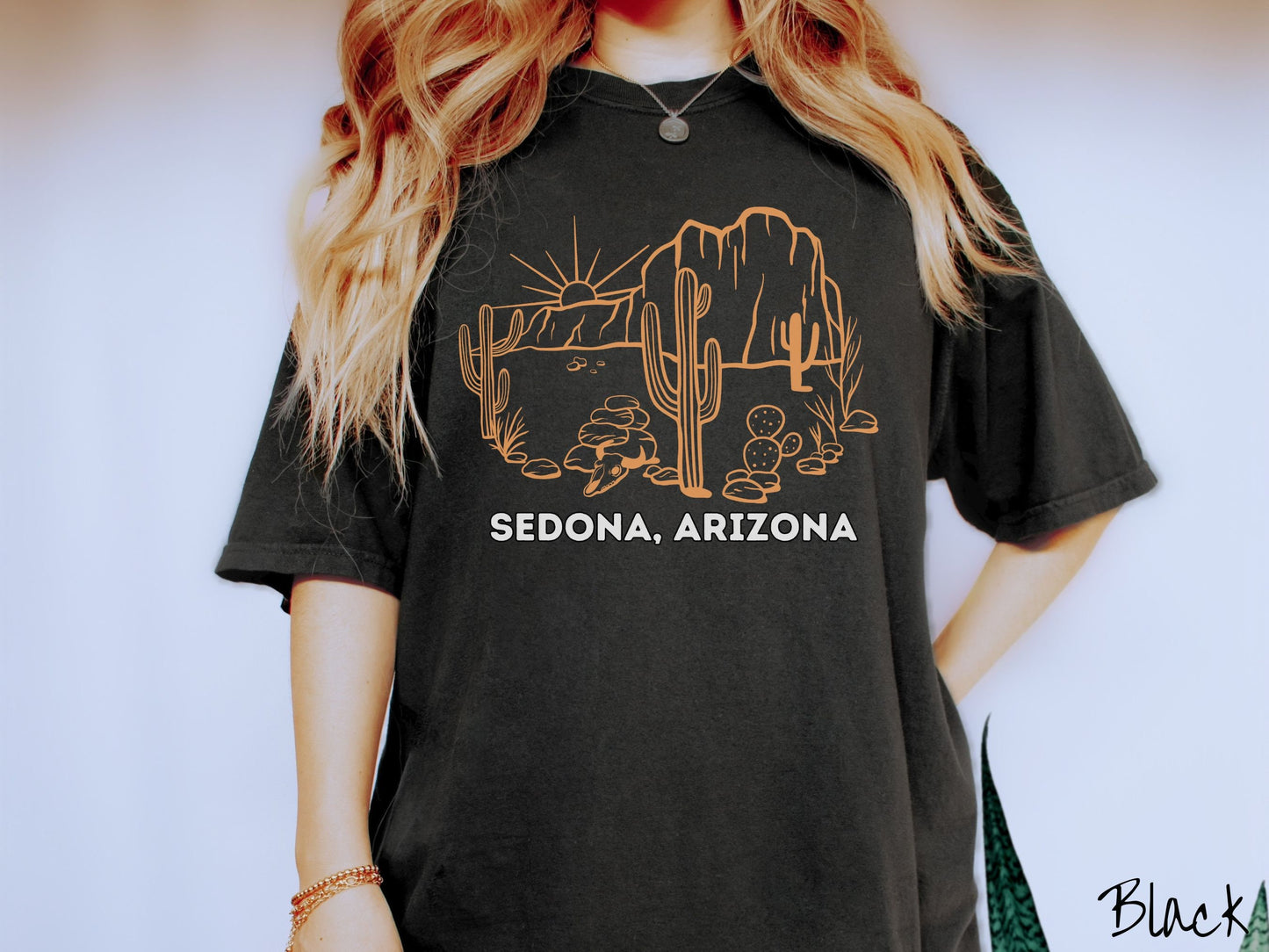A woman wearing a cute, vintage black colored Comfort Colors t-shirt with text Sedona, Arizona in white font. Above is a black outline of a desert landscape with the sun and mountains in the background, and cacti and rocks in the foreground.