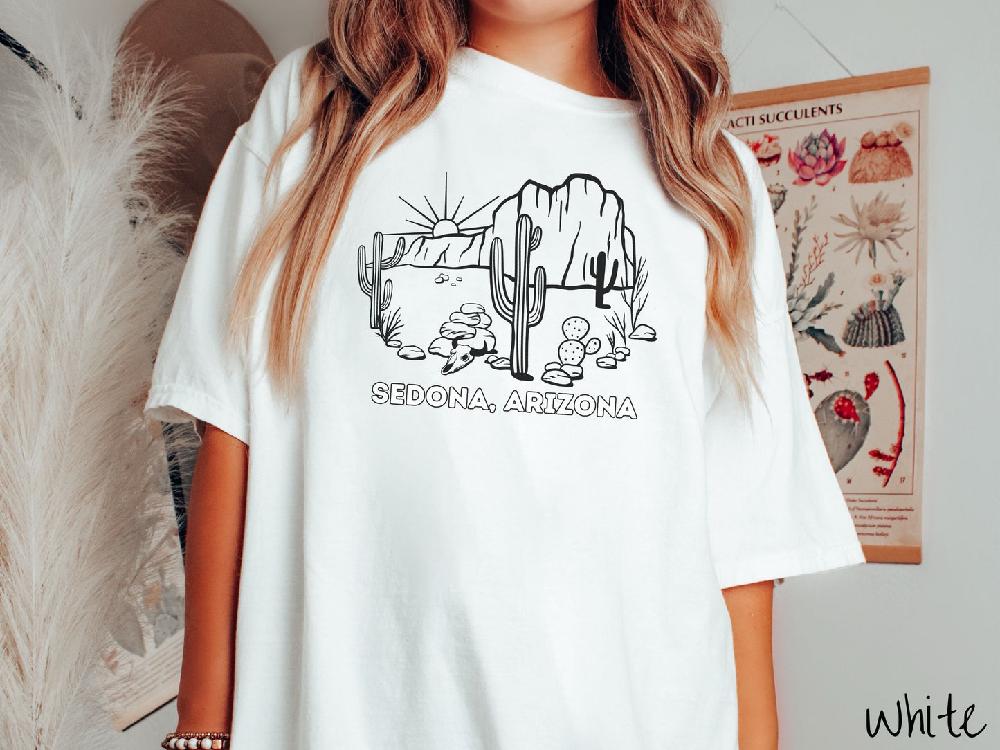A woman wearing a cute, vintage white colored Comfort Colors t-shirt with text Sedona, Arizona in white font. Above is a black outline of a desert landscape with the sun and mountains in the background, and cacti and rocks in the foreground.