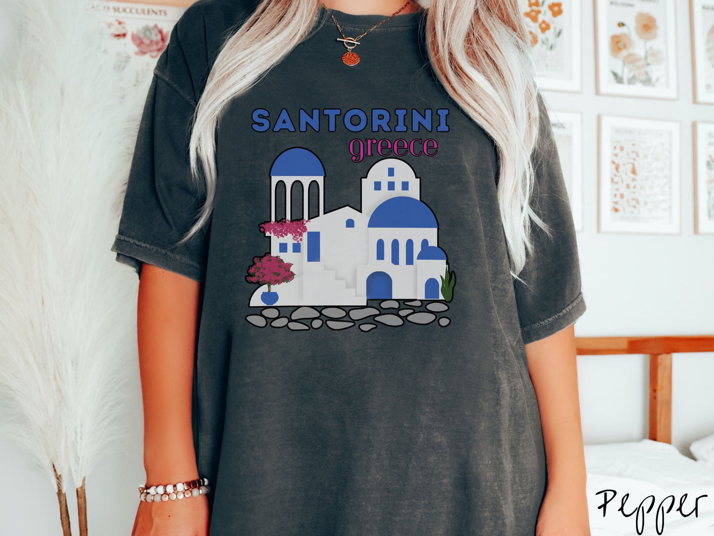 A woman wearing a cute, vintage pepper colored Comfort Colors t-shirt with text Santorini Greece in blue and pink font. Below that is a picture of the iconic cave houses with white walls and blue trim, with cobble-stone roads and red trees.