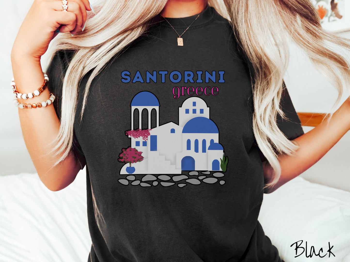 A woman wearing a cute, vintage black colored Comfort Colors t-shirt with text Santorini Greece in blue and pink font. Below that is a picture of the iconic cave houses with white walls and blue trim, with cobble-stone roads and red trees.