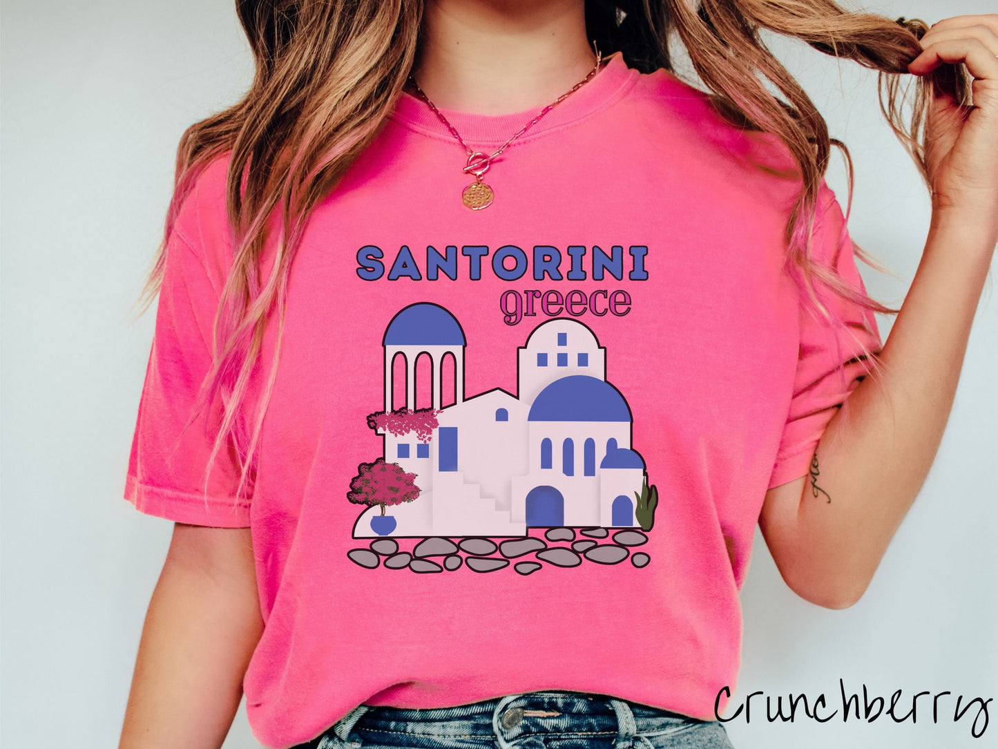 A woman wearing a cute, vintage crunchberry colored Comfort Colors t-shirt with text Santorini Greece in blue and pink font. Below that is a picture of the iconic cave houses with white walls and blue trim, with cobble-stone roads and red trees.
