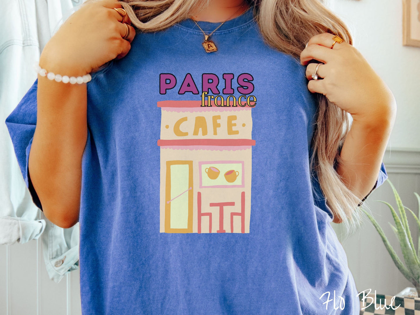 A woman wearing a cute, vintage flo blue colored Comfort Colors t-shirt with the text Paris France in purpple and yellow font. Below this is a French cafe with a coffee cup picture on the outside above a table and chairs for outside seating.