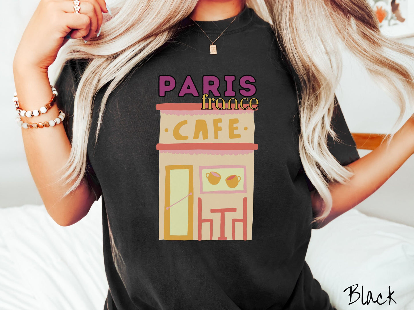 A woman wearing a cute, vintage black colored Comfort Colors t-shirt with the text Paris France in purpple and yellow font. Below this is a French cafe with a coffee cup picture on the outside above a table and chairs for outside seating.