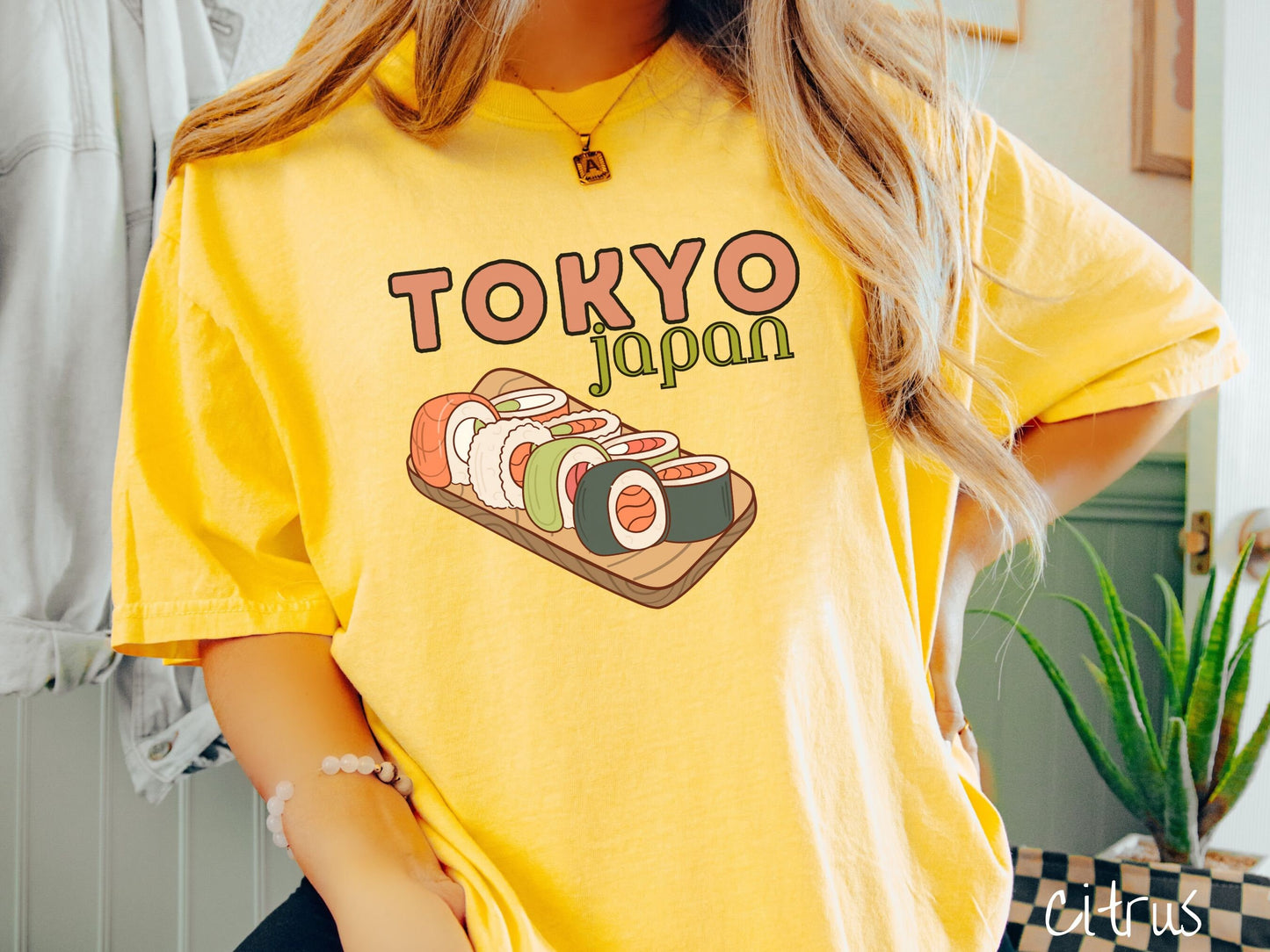 A woman wearing a vintage, citrus colored shirt with the text Tokyo Japan across the top in salmon and green font, respectively, and below is an assortment of colorful sushi rolls on a narrow wooden plate.