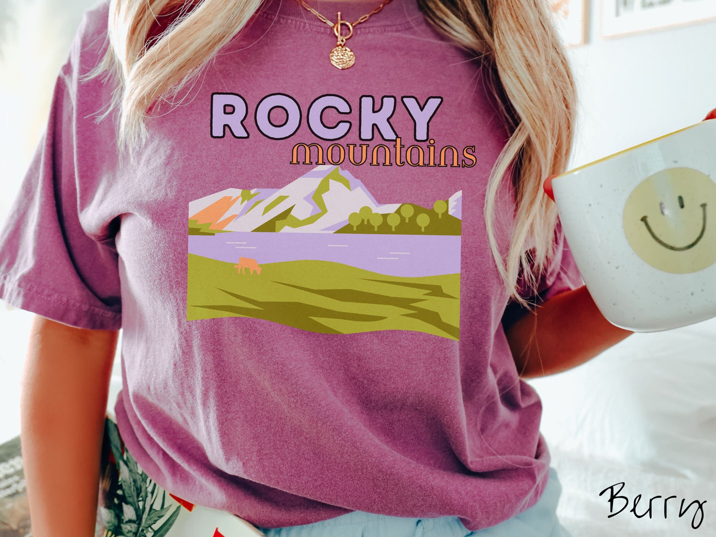 A woman wearing a vintage, berry colored shirt with the text Rocky Mountains at the top and below is a scene of a massive river running beneath snowy mountains in the distance, a lone brown cow can be seen grazing in the grass next to the water.