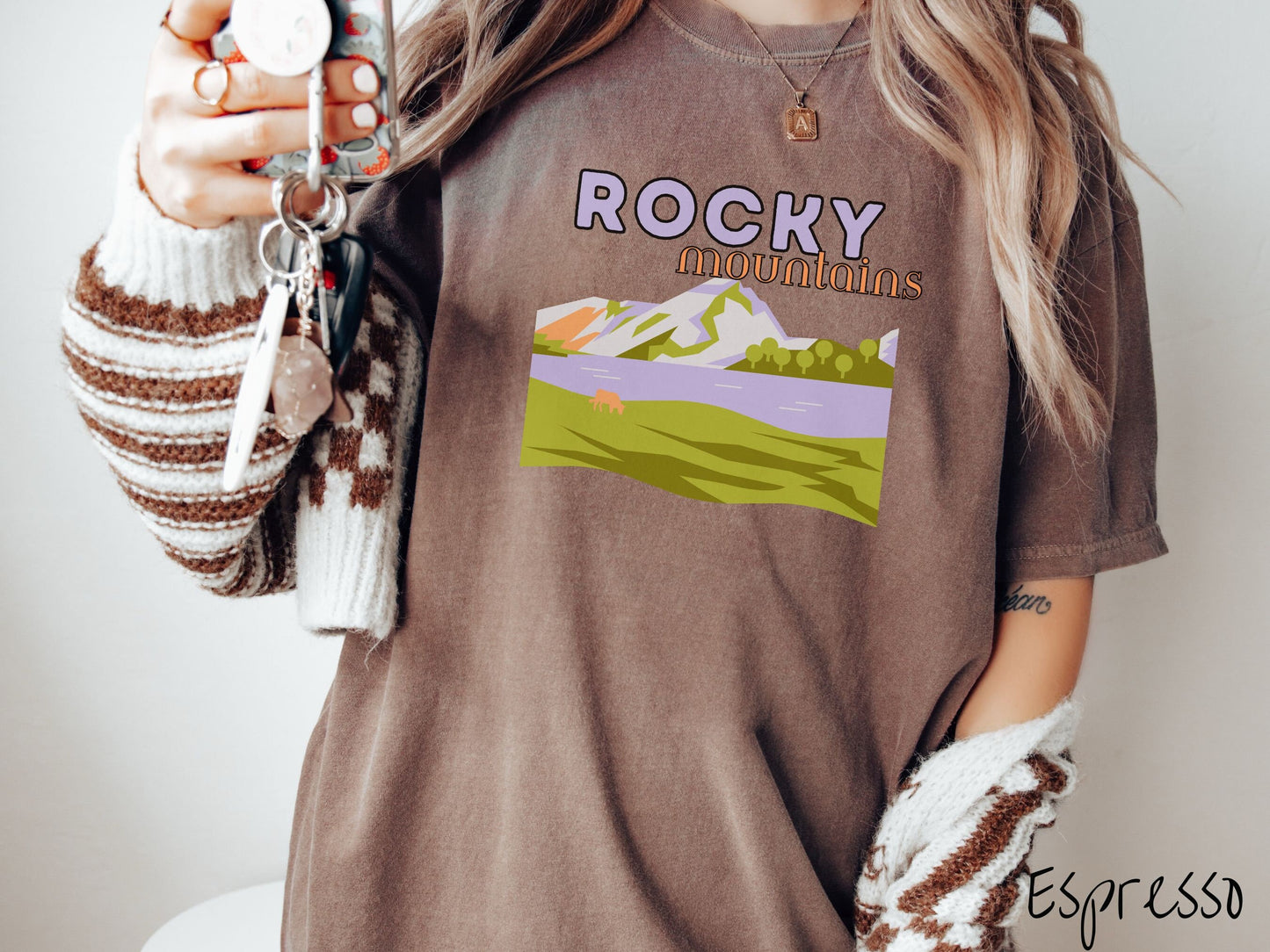 A woman wearing a vintage, espresso colored shirt with the text Rocky Mountains at the top and below is a scene of a massive river running beneath snowy mountains in the distance, a lone brown cow can be seen grazing in the grass next to the water.