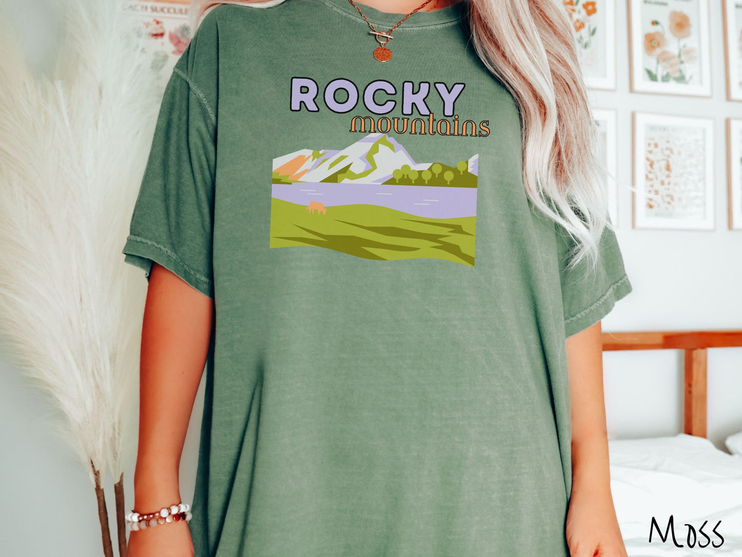 A woman wearing a vintage, moss colored shirt with the text Rocky Mountains at the top and below is a scene of a massive river running beneath snowy mountains in the distance, a lone brown cow can be seen grazing in the grass next to the water.