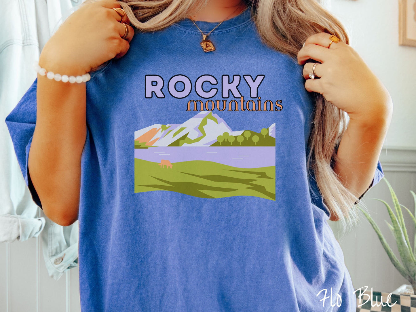 A woman wearing a vintage, flo blue colored shirt with the text Rocky Mountains at the top and below is a scene of a massive river running beneath snowy mountains in the distance, a lone brown cow can be seen grazing in the grass next to the water.