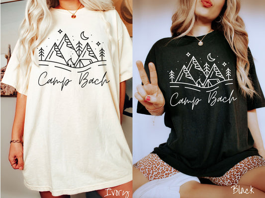 Two women wearing cute, vintage ivory and black colored Comfort Colors t-shirts with text Camp Bach in white, cursive font. Above the text are outlines of mountains, trees, stars, a tent, campfire, and a stream.