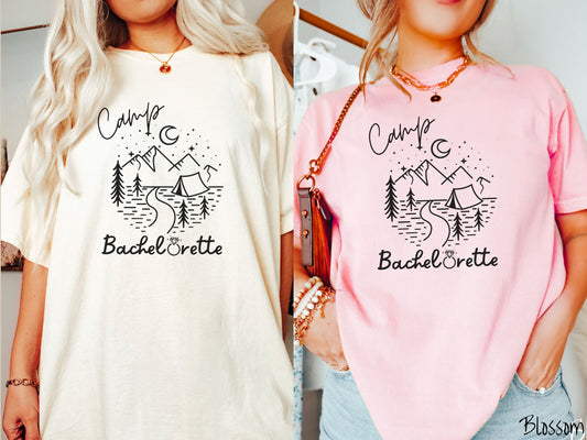 Two women wearing cute, vintage ivory and blossom colored Comfort Colors t-shirts with text Camp Bachelorette in black, cursive font. The o in Bachelorette is a diamond wedding ring. Between the text are mountains, trees, a tent, and a stream.