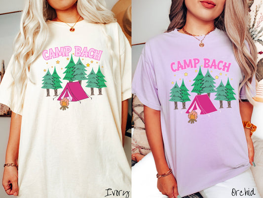 Two women wearing cute, vintage ivory and orchid colored Comfort Colors t-shirts with text Camp Bach in pink, uppercase font. Below the text is a pink tent in between green trees and in front of a campfire with stars in the sky above the trees.