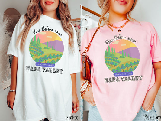 Two women wearing cute, vintage white and blossom colored Comfort Colors t-shirts with text Vino before vows and Katie’s Bachelorette Napa Valley, with a picture of a California wine vineyard with rows of trees in a meadow with purple mountains.