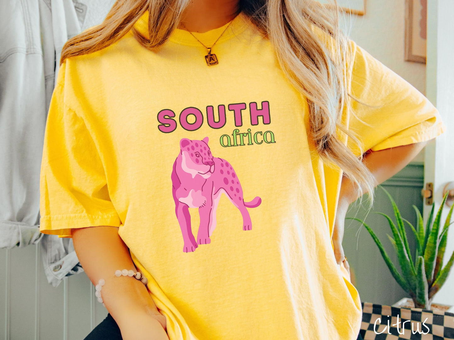A woman wearing a vintage, citrus colored shirt with the text South Africa in pink and green font, respectively, and a pink tiger underneath on the prowl.