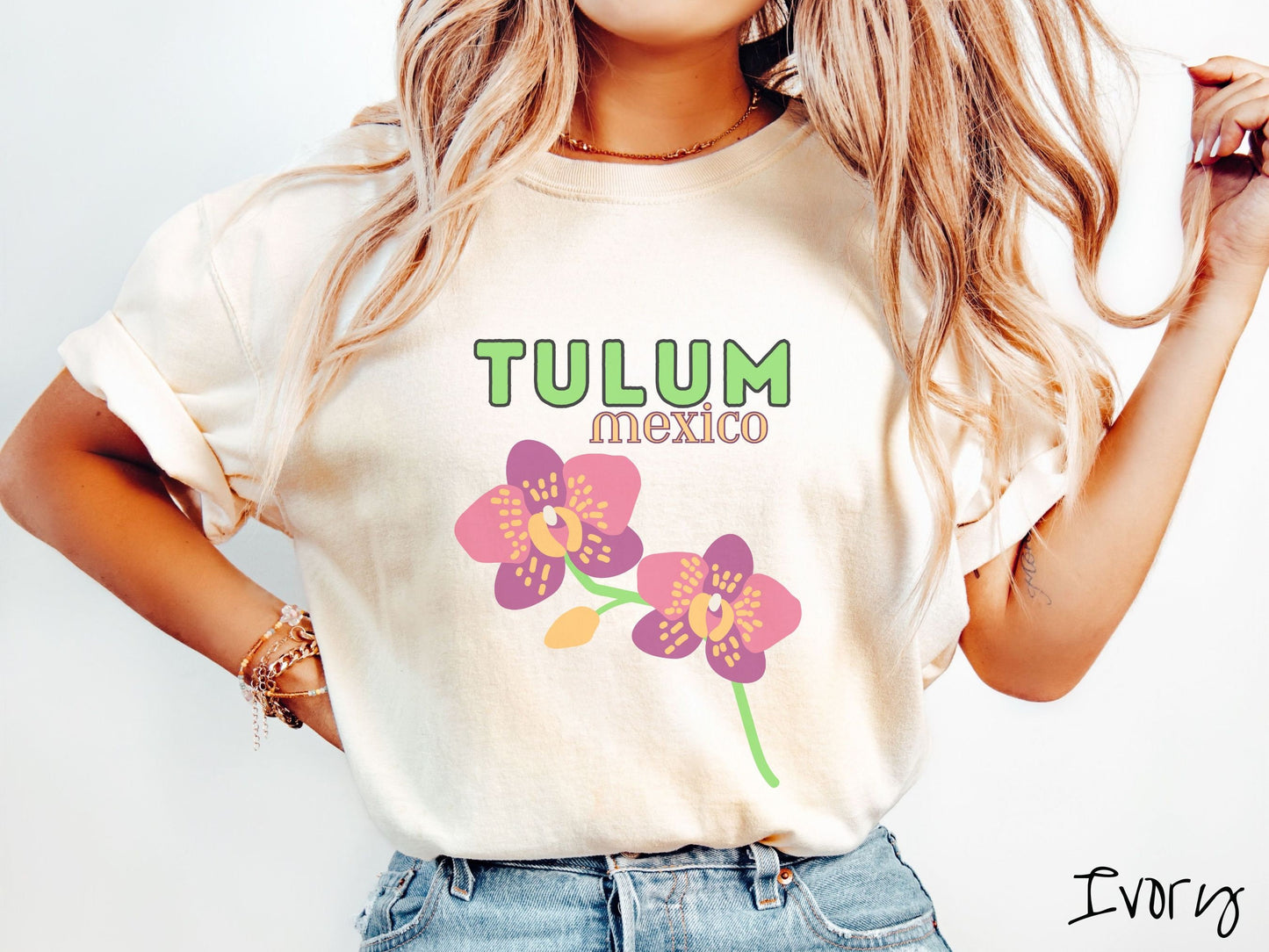 A woman wearing a vintage, ivory colored shirt with the text Tulum Mexico in green and yellow font, respectively, at the top and below it are two beautiful pink and purple flowers with green stems.